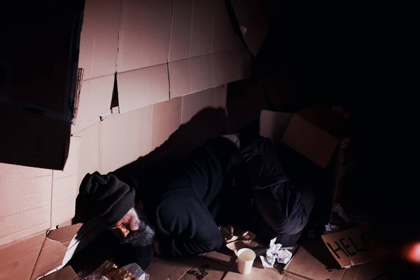 A homeless bearded man sits on boxes on street and asks for help. Need a homeless person asks for money for food and overnight. - Zdjęcie, obraz