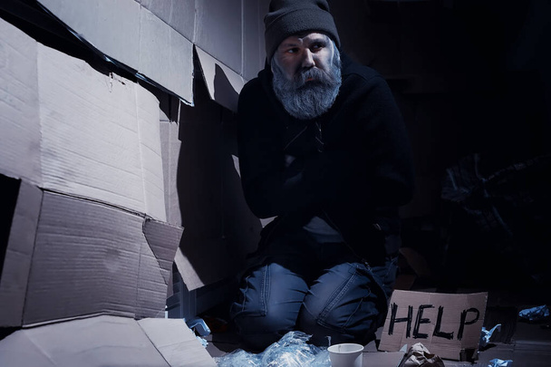 Ein bärtiger Obdachloser sitzt auf Kisten auf der Straße und bittet um Hilfe. Ein Obdachloser bittet um Geld für Essen und Übernachtung. - Foto, Bild