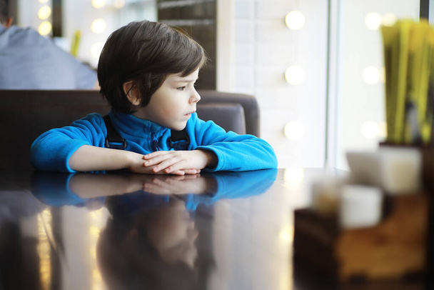 Genç bir çocuk kafenin penceresinin yanındaki masada oturur ve bir garsonun gelip sipariş vermesini bekler.. - Fotoğraf, Görsel