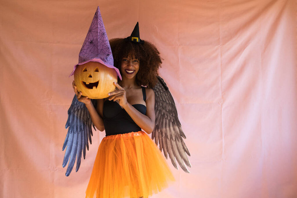 ハロウィンパーティーの魔女の衣装を着た若いアフリカ系アメリカ人女性。彼女は魔女の帽子、オレンジのスカート、黒い天使の翼を着ています。女性はカボチャと遊んでいる間にカメラで笑顔. - 写真・画像
