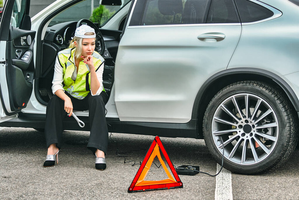 sad woman in emergency vest sits near the car. automobile breakdown - Фото, изображение