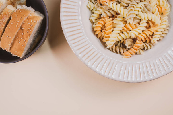 A plate of pasta twists and a bowl of sliced bread on a beige background with free space for text - 写真・画像