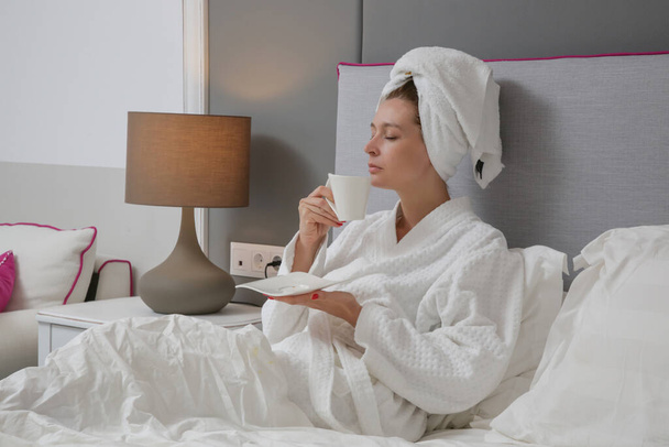 Nuori kaunis nainen kylpytakissa juo kuumaa kahvia sängyssä ja nauttii hyvää huomenta. Rentoudu ja huoleton päivän alku. - Valokuva, kuva