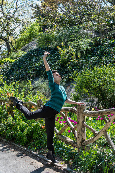 Stretching arms and legs, a young handsome guy is dancing in the garden. There are many flowers in the background - Photo, Image