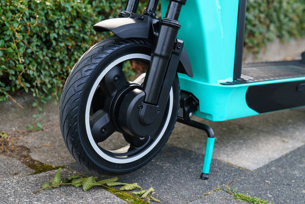 Roda dianteira de uma scooter elétrica close-up. Uma moldura azul-turquesa leve em um suporte fica na calçada. - Foto, Imagem
