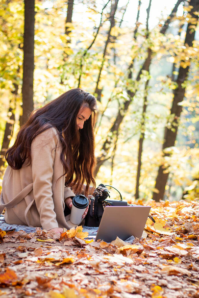 A brunette woman is sitting in an autumn park with a laptop and a cup of coffee. Student. Teacher. Online education. - Φωτογραφία, εικόνα