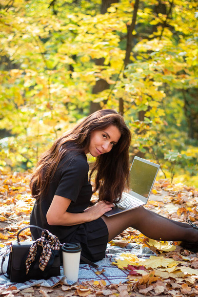 A brunette woman is sitting in an autumn park with a laptop and a cup of coffee. Student. Teacher. Online education. - Фото, зображення