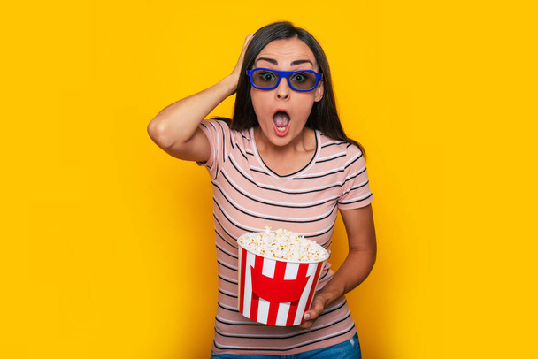 Excited beautiful modern young brunette woman in casual wear and 3d cinema glasses and big basket of popcorn is posing while watching some film in cinema  - Foto, Imagen