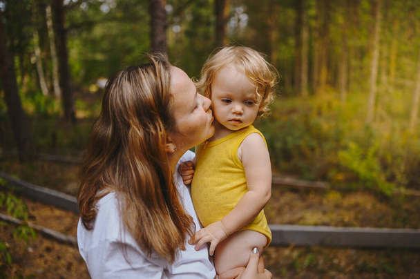 Annesinin kollarında bukleleri olan küçük tatlı bebek sarışın kız. Anne ve kızı yazın kırsal alanda ön bahçede birlikte vakit geçiriyorlar. Sağlıklı mutlu aile çocukluğu kavramı. - Fotoğraf, Görsel