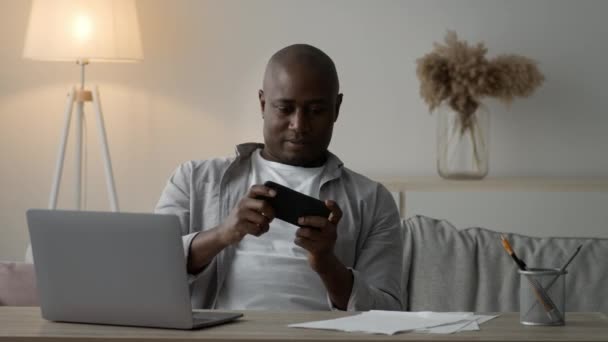 Cheerful Black Man Using Smartphone Playing Game Online Sitting Indoors - Footage, Video