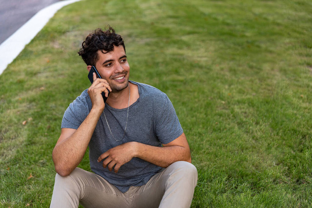 Joven latino sentado en la hierba hablando por teléfono móvil. - Foto, imagen