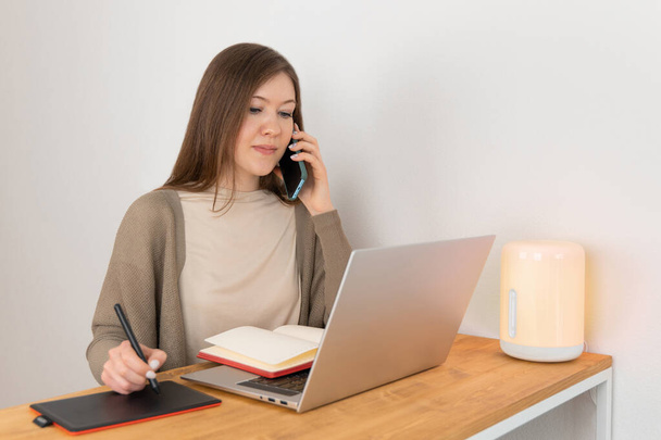 Handsome caucasian woman in beige t-shirt and grey cardigan calling on mobile phone and looking into laptop display. Attractive girl makes handwritten notes on a graphic tablet. Home office concept. - Fotografie, Obrázek