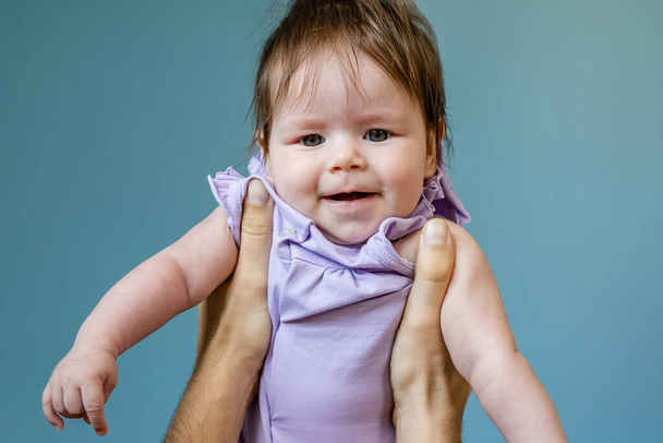 Manos de hombre desconocido padre sosteniendo a su hija bebé niña en el aire contra el niño de la pared azul usando vestido púrpura en el concepto de paternidad y paternidad del día de verano creciendo - Foto, Imagen