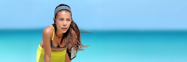 Tired running Asian woman listening to music or podcast with wireless earbuds wearing phone armband holder during beach run sweating on summer workout. Heat stroke breathing fitness girl banner. - Photo, Image