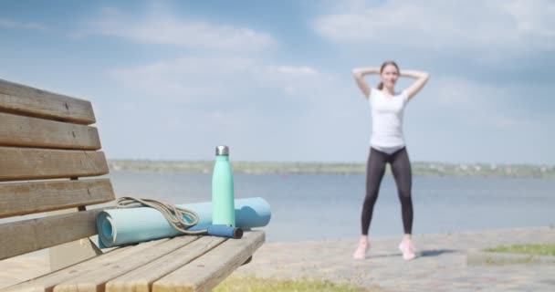 Αθλητική νεαρή γυναίκα προπόνηση κοντά στο ποτάμι, επικεντρωθεί στην άλμα σχοινί, στρώμα γιόγκα και μπουκάλι νερό στον πάγκο - Πλάνα, βίντεο