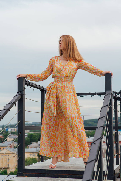 Beautiful young woman in long leopard dress standing with bare feet on the stairs on the top of the roof of a building. - Foto, Bild