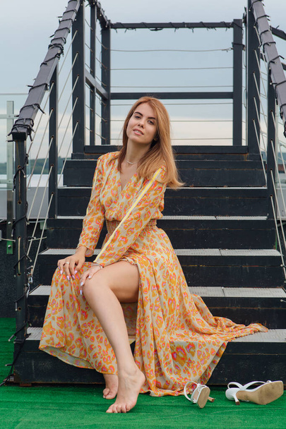 Uzun leopar desenli elbiseli güzel bir kadın merdivenlerde çıplak ayakla bir binanın çatısında oturuyor.. - Fotoğraf, Görsel