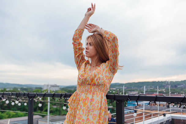 Beautiful young woman in long leopard dress standing on the stairs on the top of the roof of a building. - Foto, afbeelding