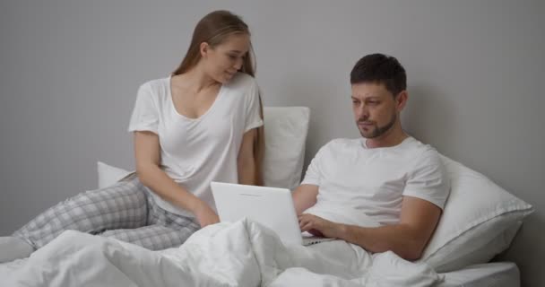 Mujer coquetea con su marido en la cama, él está ocupado y utiliza el ordenador portátil, mujer se enoja - Metraje, vídeo