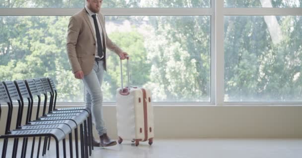 Hombre con maleta y teléfono móvil esperando su vuelo en el aeropuerto - Metraje, vídeo