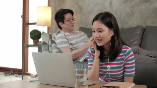 Aasian nainen käyttää kannettavaa tietokonetta online-liiketoimintaa, verkkokaupan chat, ja internet-oppiminen, hänen veljensä sohvalla takana. Molemmat sisarukset työskentelevät kotona, koska he joutuvat karanteeniin COVID-19. - Materiaali, video