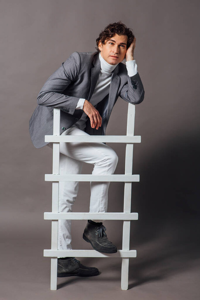 Fashion portrait of a tall handsome man dressed in white turtleneck, white jeans and grey jacket posing on the grey background with white ladder - 写真・画像