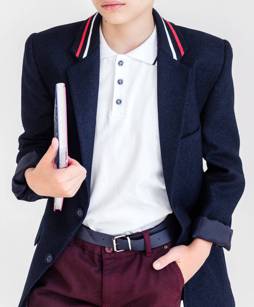 Photo A schoolboy stands on a white background in a school uniform - trousers, a jacket and a shirt. A boy holding a textbook under his arm - Foto, afbeelding
