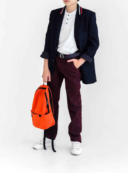 Photo of A schoolboy with an orange bag in his hands stands on a white background in a school uniform - trousers, a jacket and a shirt. - Fotó, kép