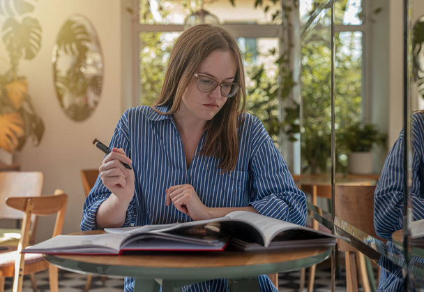 Student in eyeglasses in modern cafe with books and syllabus, studying in trendy coffee shop with day light - Foto, afbeelding