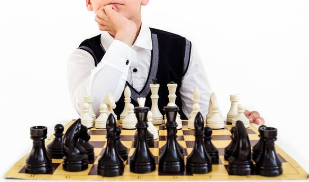 Ένα αγόρι με λευκό πουκάμισο και μαύρο γιλέκο παίζει σκάκι και σκέφτεται την επόμενη κίνηση. Φωτογραφία - Φωτογραφία, εικόνα