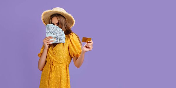 Λευκή γυναίκα 20 ετών με ψάθινο καπέλο που κρατά πιστωτική κάρτα και οπαδός του δολαρίου μετρητά που απομονώνονται σε μωβ φόντο - Φωτογραφία, εικόνα