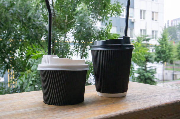 カフェ、コーヒーショップ、レストランの木製テーブルの上に晴れた日の夏に屋外に蓋付きの2つの黒い紙のコーヒーカップ - 写真・画像