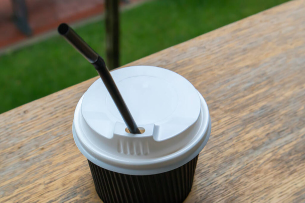 Ένα μαύρο χάρτινο φλιτζάνι καφέ με καπάκι σε εξωτερικούς χώρους το καλοκαίρι σε ηλιόλουστο καιρό σε ένα ξύλινο τραπέζι ενός καφέ, καφετέρια ή εστιατόριο - Φωτογραφία, εικόνα