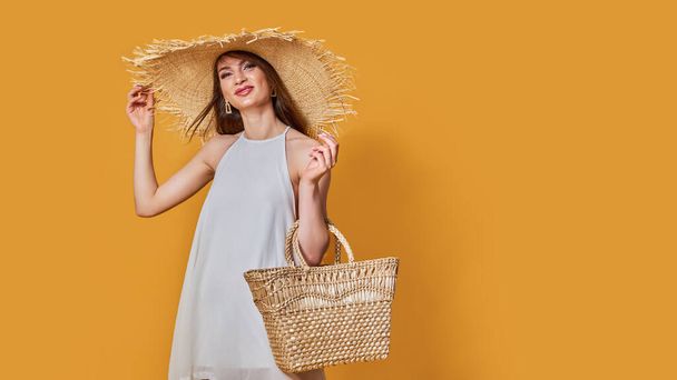Pretty glamorous girl touching her straw hat. Studio portrait of joyful young woman with summer bag. - Foto, Imagem