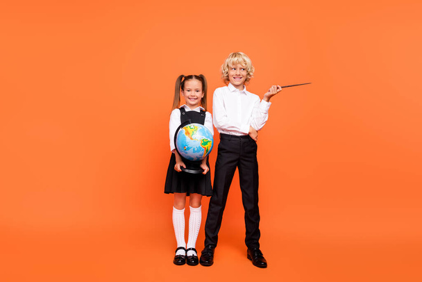 Photo of two positive curious children hold globe pointer empty space wear uniform isolated orange color background - Fotó, kép