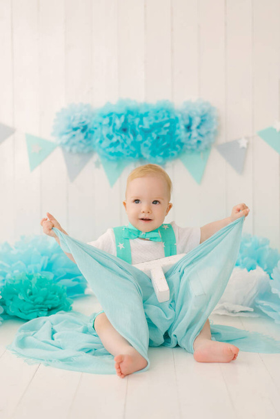 Piękny, roczny chłopiec w turkusowych ubraniach bawi się kawałkiem niebieskiej tkaniny. - Zdjęcie, obraz