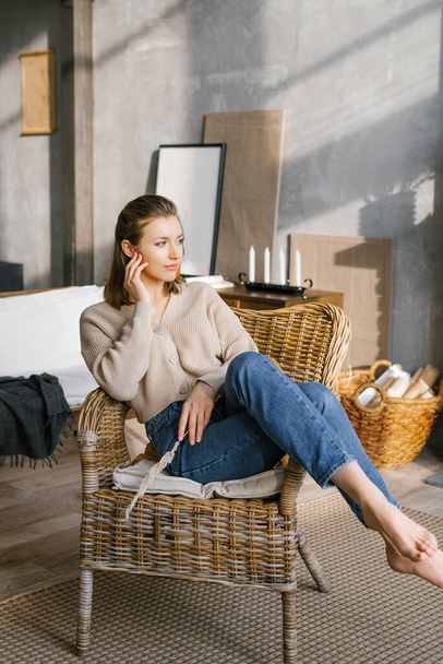 Красивая молодая женщина в джинсах и свитер сидит на плетеном стуле у камина дома и мечтает - Фото, изображение