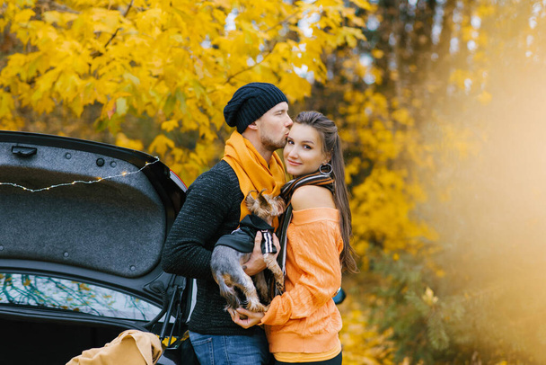 Een jongen en een verliefd meisje omhelzen in een herfstpark, een jongen houdt een schattige hond in zijn armen en kust zijn favoriete meisje - Foto, afbeelding