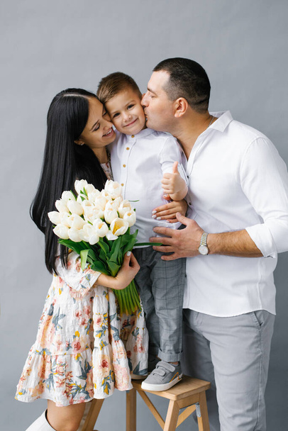A happy young family. Beautiful mother and father kiss their child . Parents, A portrait of mom, dad and a smiling child isolated on a gray background. - Photo, Image