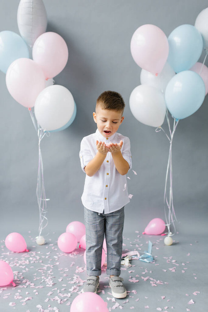 Wszystkiego najlepszego, dziecko. Zdjęcie uroczego małego chłopca dmuchającego na konfetti na tle balonów - Zdjęcie, obraz