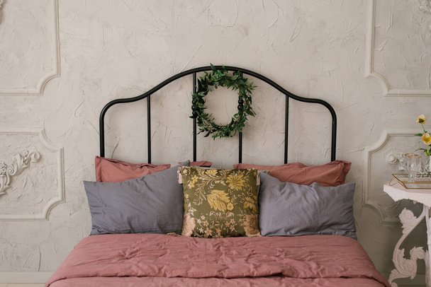Een bed met roze stoffig beddengoed en grijze kussens in Scandinavische of klassieke stijl. Een krans van bladeren hangt op het bed - Foto, afbeelding