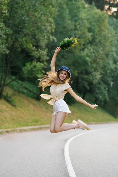 A happy girl in white shorts and a yellow blouse jumps up with a bouquet of flowers, standing on the road near the forest - Photo, Image