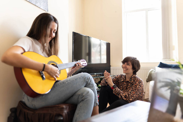 Приваблива молода жінка грає пісню зі своєю гітарою, в той час як її друг слухає і оживляє з клаптиками і сміється
 - Фото, зображення