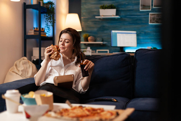 Счастливая женщина ест вкусные бургеры, расслабляясь на диване, смотря комедийный фильм - Фото, изображение