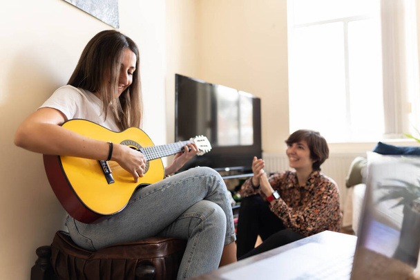 Φωτογραφία μιας ελκυστικής νεαρής γυναίκας που παίζει κιθάρα ενώ η φίλη της την ακούει και ζωντανεύει με χειροκροτήματα και γέλια. - Φωτογραφία, εικόνα