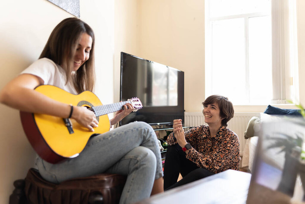 Приваблива молода жінка грає пісню зі своєю гітарою, в той час як її друг слухає і оживляє з клаптиками і сміється
 - Фото, зображення