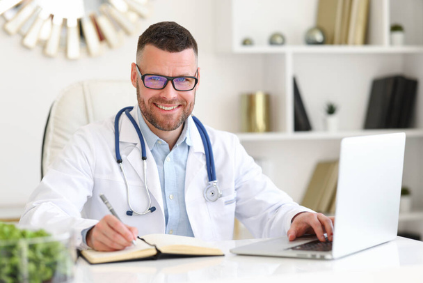 Fröhlich-freundlicher Arzt in weißem Mantel mit Stethoskop auf den Schultern, der lächelnd in die Kamera blickt, während er im Krankenhaus am Laptop arbeitet, männlicher Arzt sitzt an seinem Arbeitsplatz  - Foto, Bild
