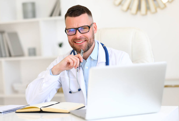 Cheerful friendly male doctor wearing white coat with stethoscope on shoulders looking at camera with smile while working on a laptop in hospital, male physician sitting at his workplace  - Foto, afbeelding