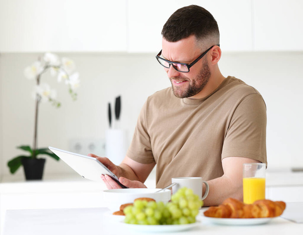 Jonge glimlachende knappe man met een bril die op afstand op tablet werkt terwijl hij thuis in de moderne keuken zit, mannelijke freelancer die gezond ontbijt eet en naar de computer kijkt - Foto, afbeelding