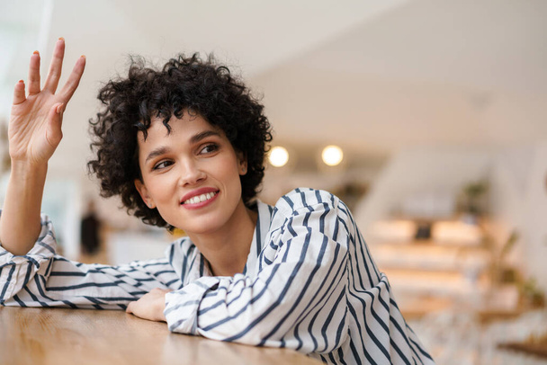 Beautiful happy curly woman smiling and waving hand while sitting in cafe - Photo, Image